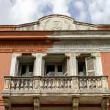 Da qui riusciamo a scorgere meglio i decori della facciata. Nella parte centrale spicca il balcone in pietra, oltre il quale intravediamo quattro bianche colonne scanalate...